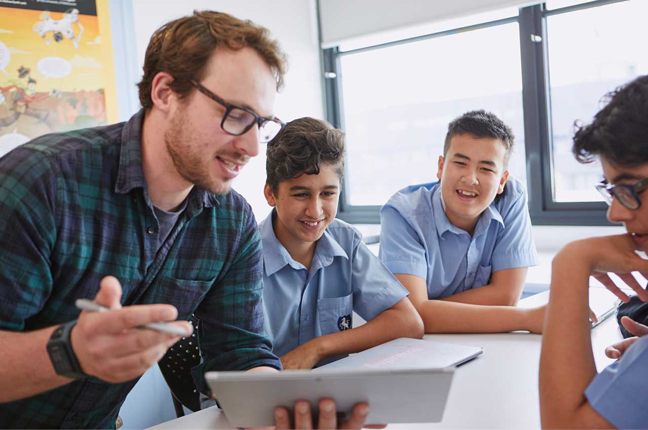 A teacher is showing something on a tablet to three students. The students are leaning in to see and are enjoying themselves