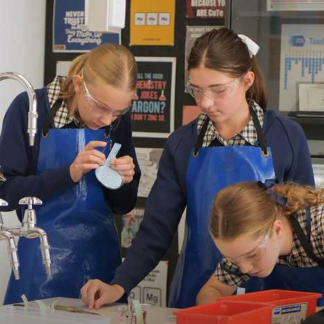 Three students in school uniforms wearing safety goggles and blue aprons working on a science experiment at a lab bench.