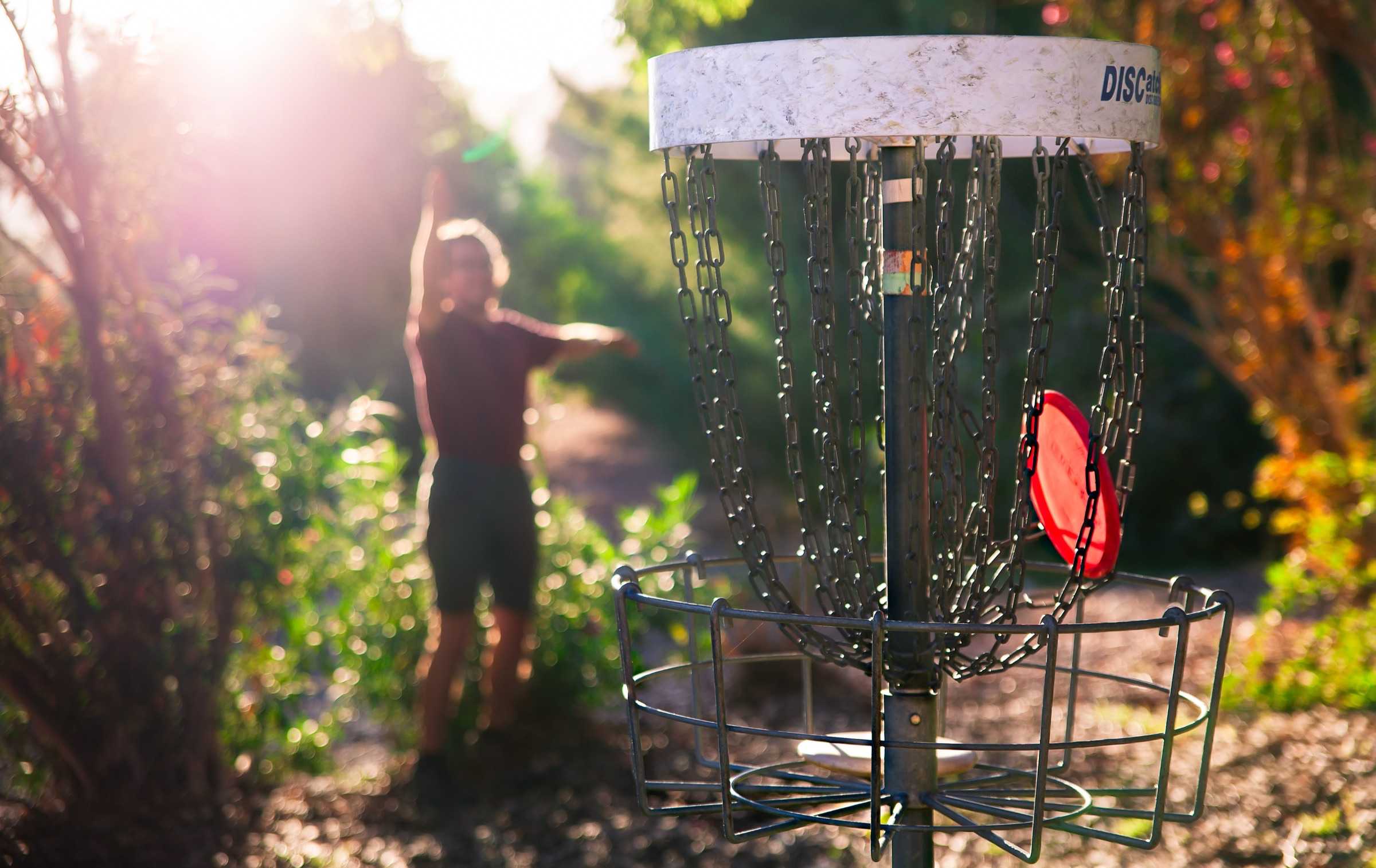 A disc golfer throwing a frisbee toward a basket in a wooded outdoor course.