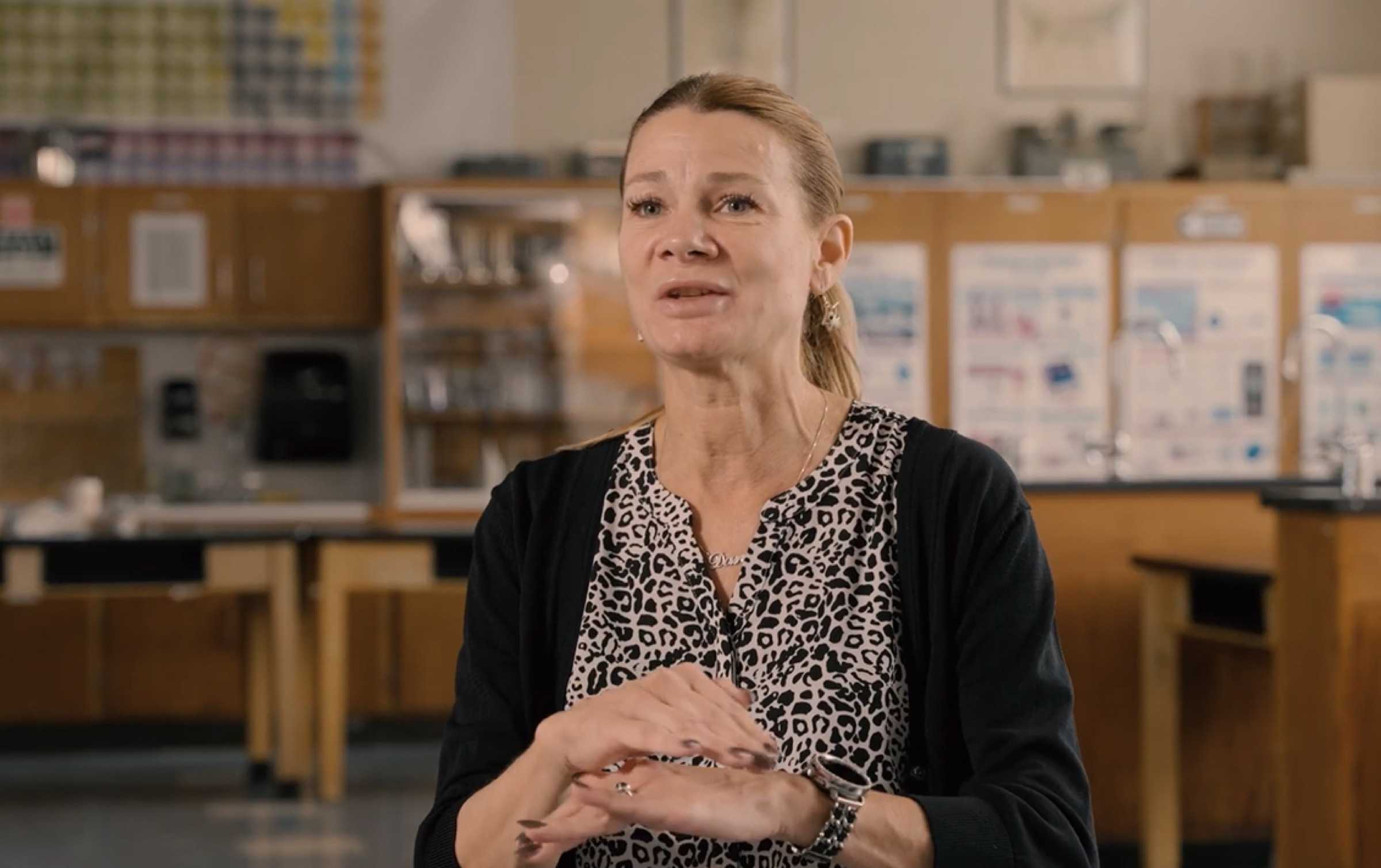 A teacher speaking in a middle school science classroom with lab tables and equipment in the background.