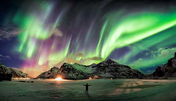 A wide-angle show of a person standing in a frozen plain, with mountains and an aurora in the sky behind them