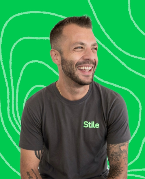 Mark Picardo smiling, wearing a Stile t-shirt, in front of a green background with abstract line patterns.
