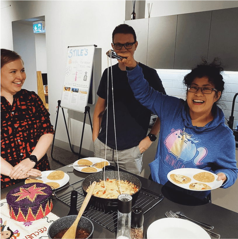 Three team members in the office kitchen, cooking together. One is pulling a very long string of cheese out of a pan