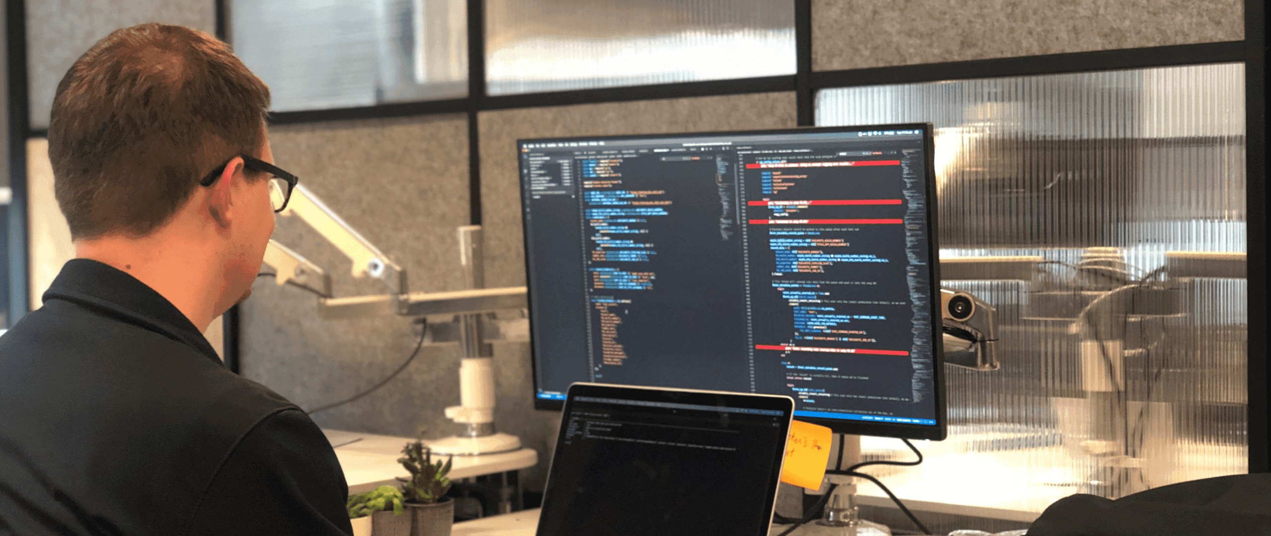 A software engineer at a workstation with a laptop and a wide external monitor