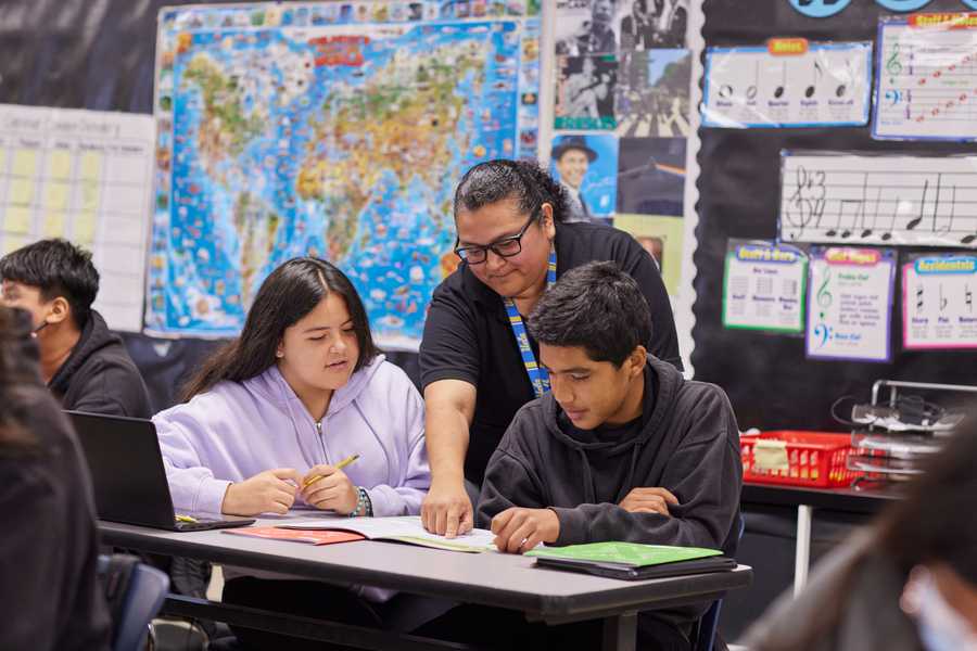 A teacher leans over and points to a students work, as another student looks on