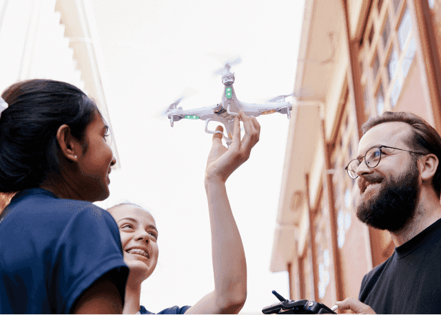 A student holds up a drone, with another student and an adult looking on