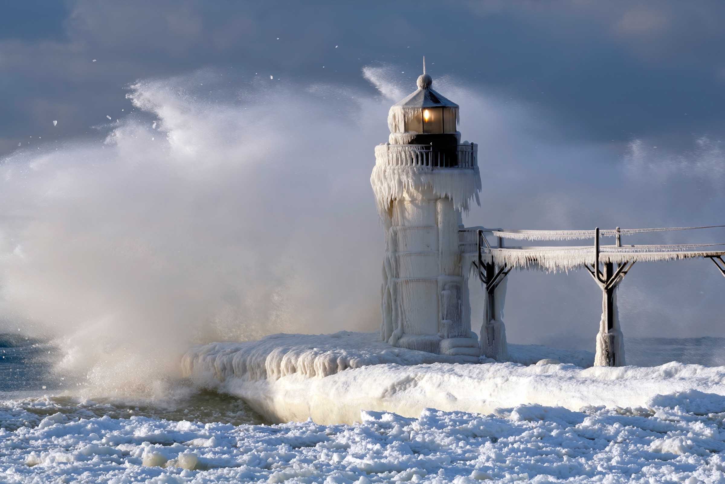 A lighthouse encased in ice with waves crashing around it during a winter storm.