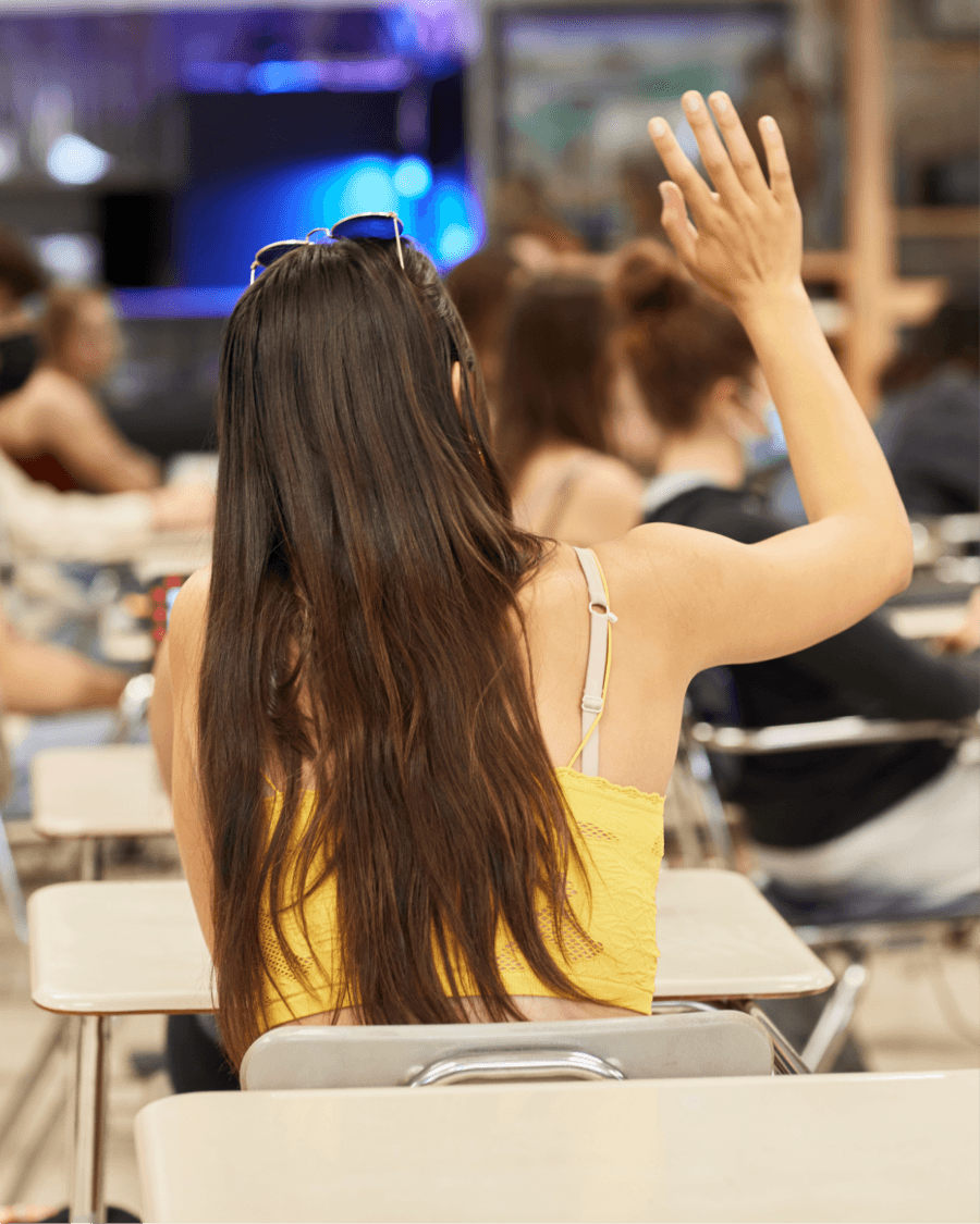 A student at a desk raising her hand, seen from behind